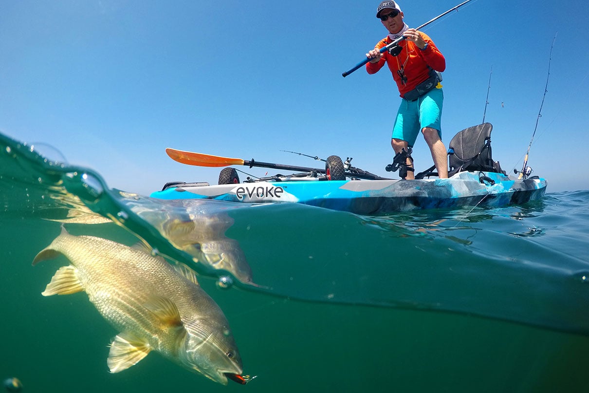 man stands on the deck of his sit-on-top fishing kayak and reels in a catch while fishing in saltwater