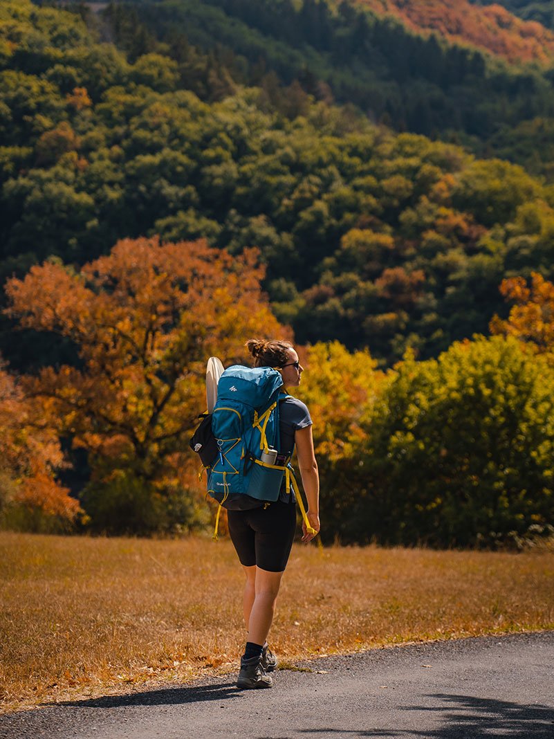 woman hikes down a roadside in front of hills with a packraft and paddle in her pack