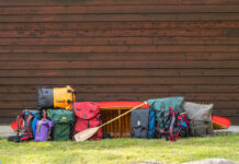 a selection of canoe packs and barrel harnesses in front of a red wood-canvas canoe and wood slat wall