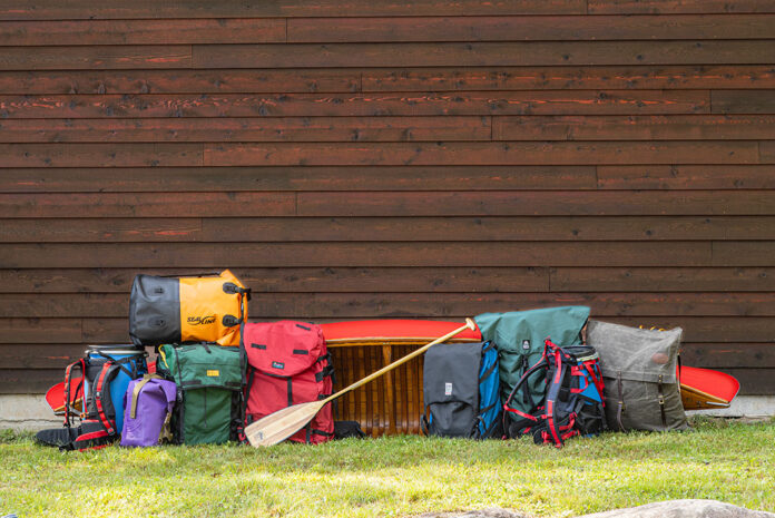 a selection of canoe packs and barrel harnesses in front of a red wood-canvas canoe and wood slat wall