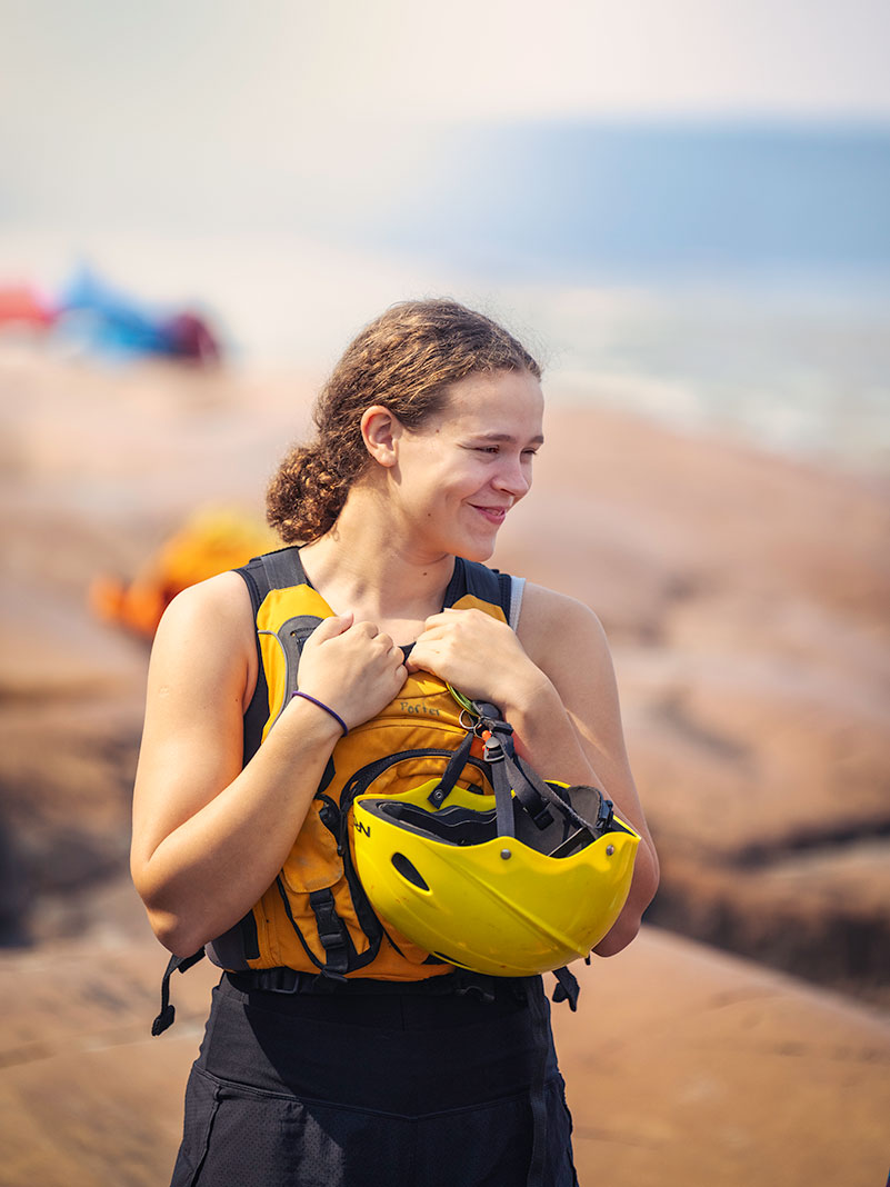 young woman paddler stands beside the Slave River while holding a yellow whitewater helmet