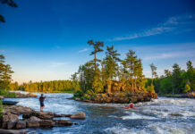 a person on the bank watches while a whitewater kayaker paddles in rapids in front of a small river island