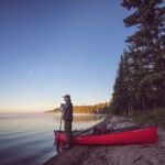 man stands at lake's edge holding a paddle beside a tripping canoe at dawn