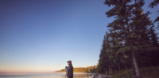 man stands at lake's edge holding a paddle beside a tripping canoe at dawn