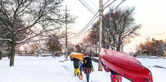 three people carry Christmas gifts and canoes along a snowy sidewalk