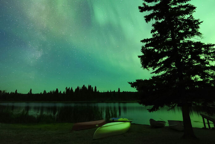 an overturned canoe sits on the bank at a campsite under a vivid night sky with northern lights