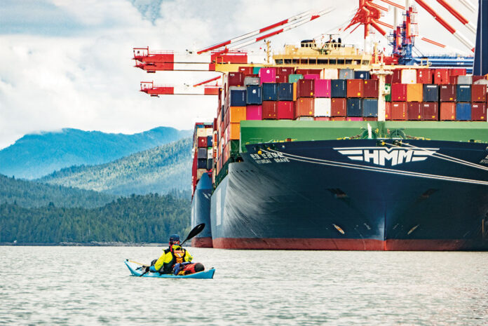man paddles a kayak in front of a large container ship in British Columbia