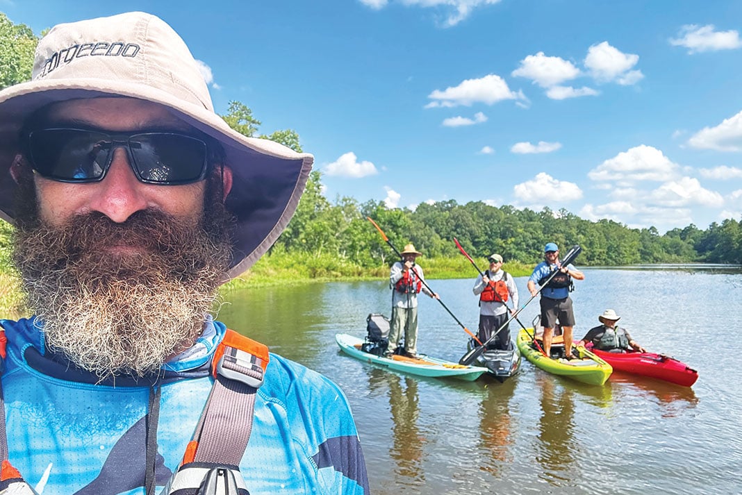 man in bucket hat and PFD takes photo with three kayak anglers posing on boats in the background