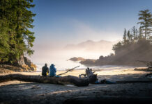 two people sit on a log on a misty beach looking out at the ocean