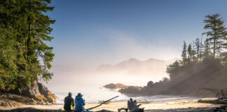 two people sit on a log on a misty beach looking out at the ocean