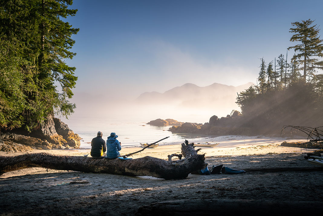 two people sit on a log on a misty beach looking out at the ocean