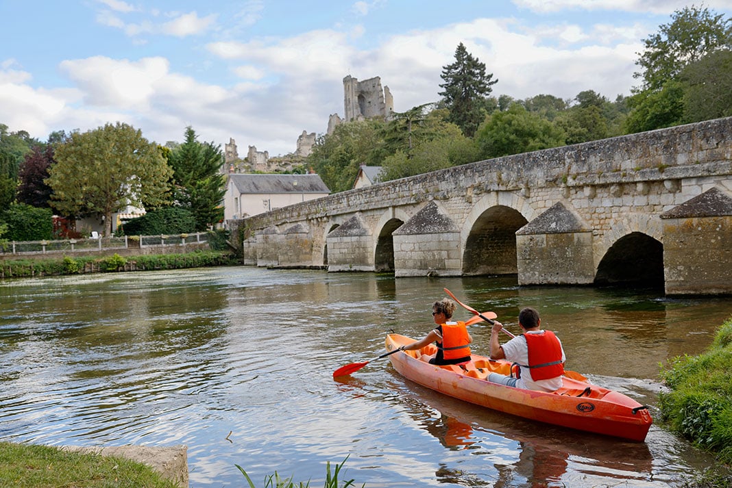 two people paddle a tandem kayak near an old bridge in France