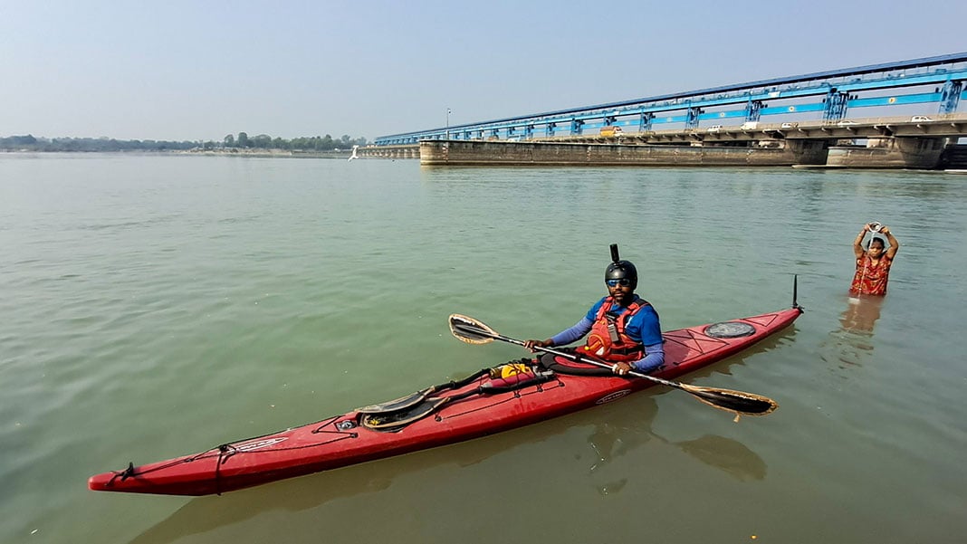 Man poses in touring kayak in India's Ganges River while woman anoints herself with water in the background