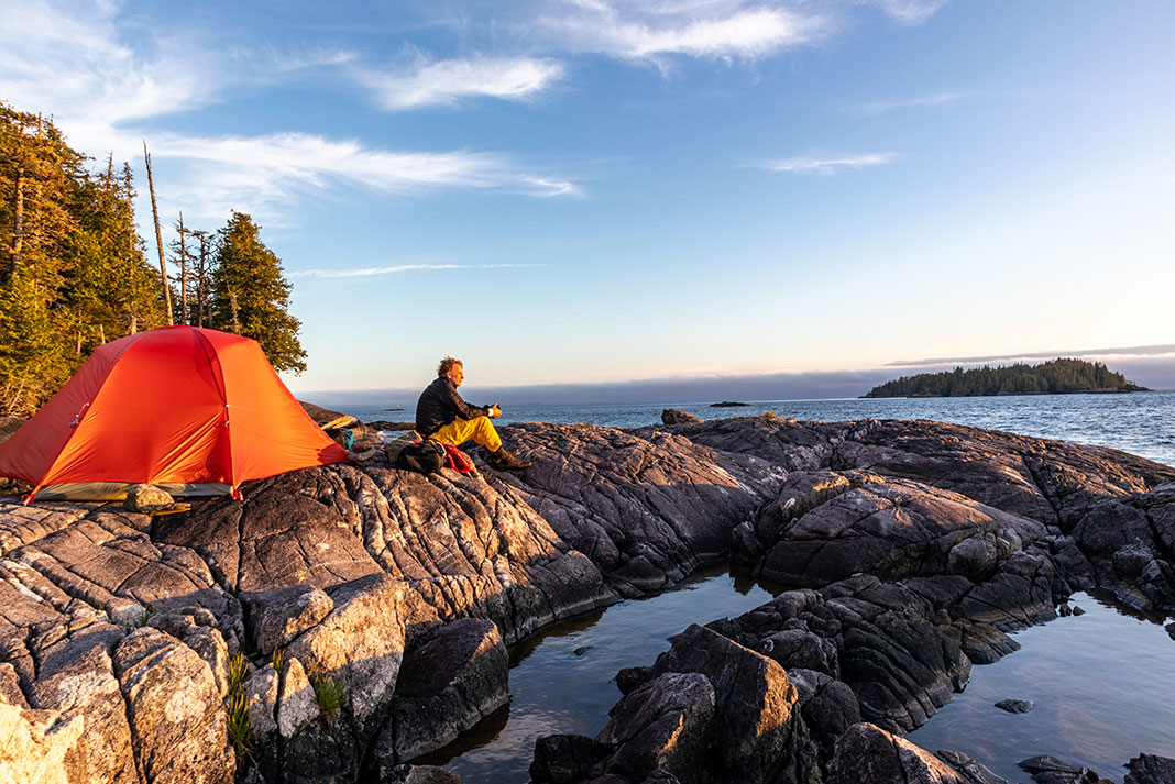 man sits on rocky West Coast shoreline near red tent and looks out over the Hecate Strait