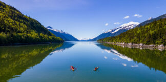 two kayakers sit on placid waters surrounded by mountains while on a summer paddling trip in Alaska