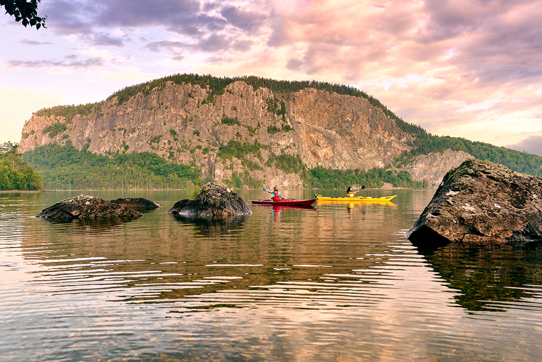 two kayakers paddle past a rocky bluff while on a summer paddling trip in Maine