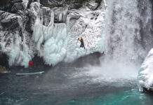 a photographer stands on his inflatable paddleboard, taking a photo of a jumping man who has been ice climbing by a waterfall