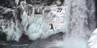 a photographer stands on his inflatable paddleboard, taking a photo of a jumping man who has been ice climbing by a waterfall