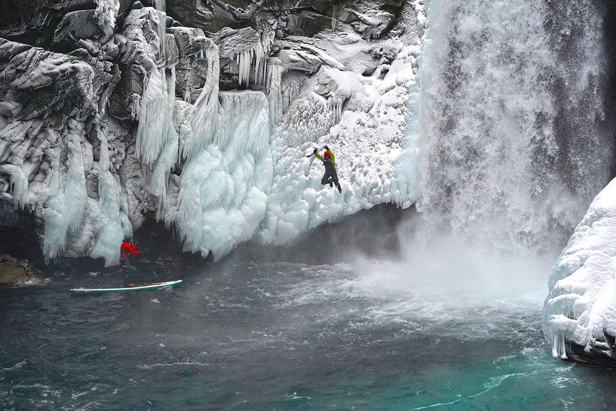 a photographer stands on his inflatable paddleboard, taking a photo of a jumping man who has been ice climbing by a waterfall