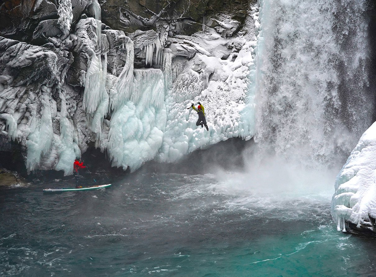 a photographer stands on his inflatable paddleboard, taking a photo of a jumping man who has been ice climbing by a waterfall