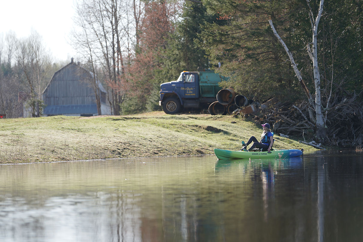 a woman pedals a sit-on-top kayak past a farm scene