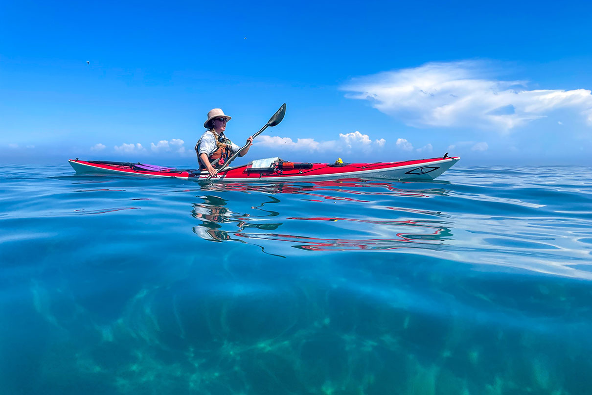 woman paddles red sea kayak in the sun on blue clear water with blue sky