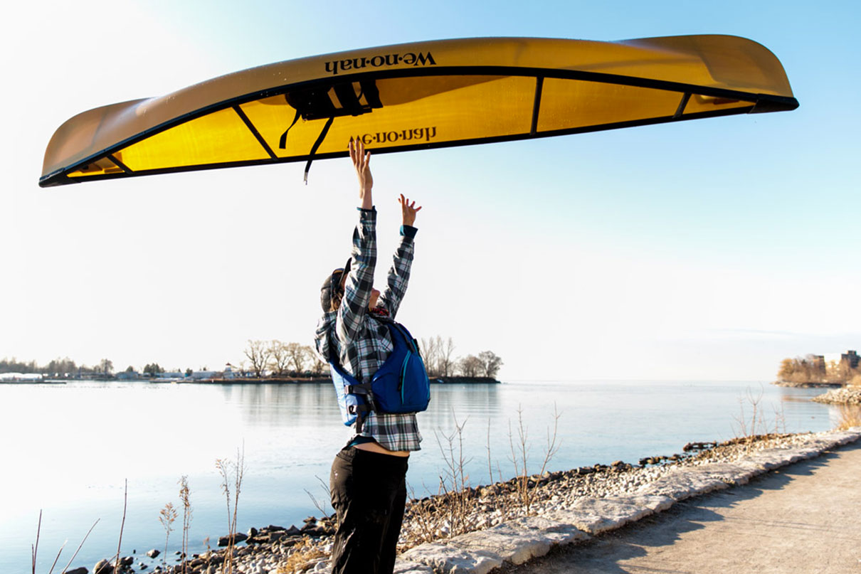 woman throws yellow ultralight canoe into the air over her head at a winter riverbank