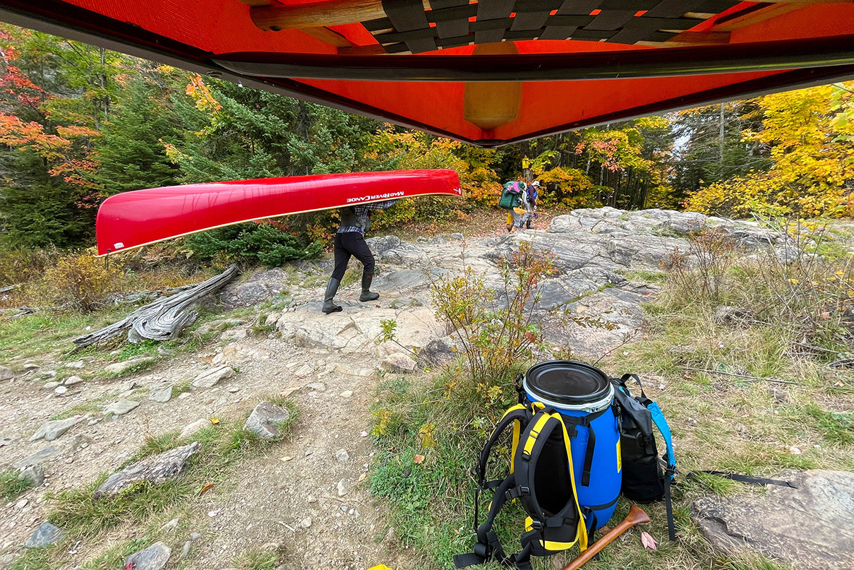 view from the vantage point of a person portaging a red canoe, with other person portaging in the background and canoe packs on the ground nearby