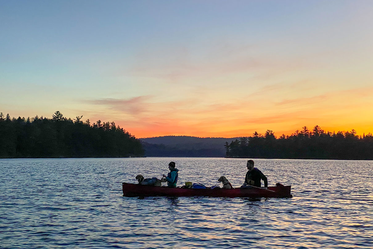 two people with two dogs paddle a loaded tripping canoe at sunset