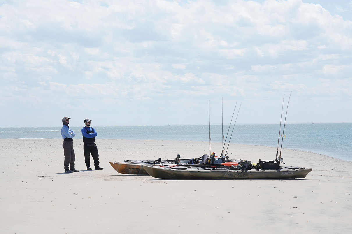 two men wearing fishing gear stand beside a pair of rigged up fishing kayaks on a white sandy beach