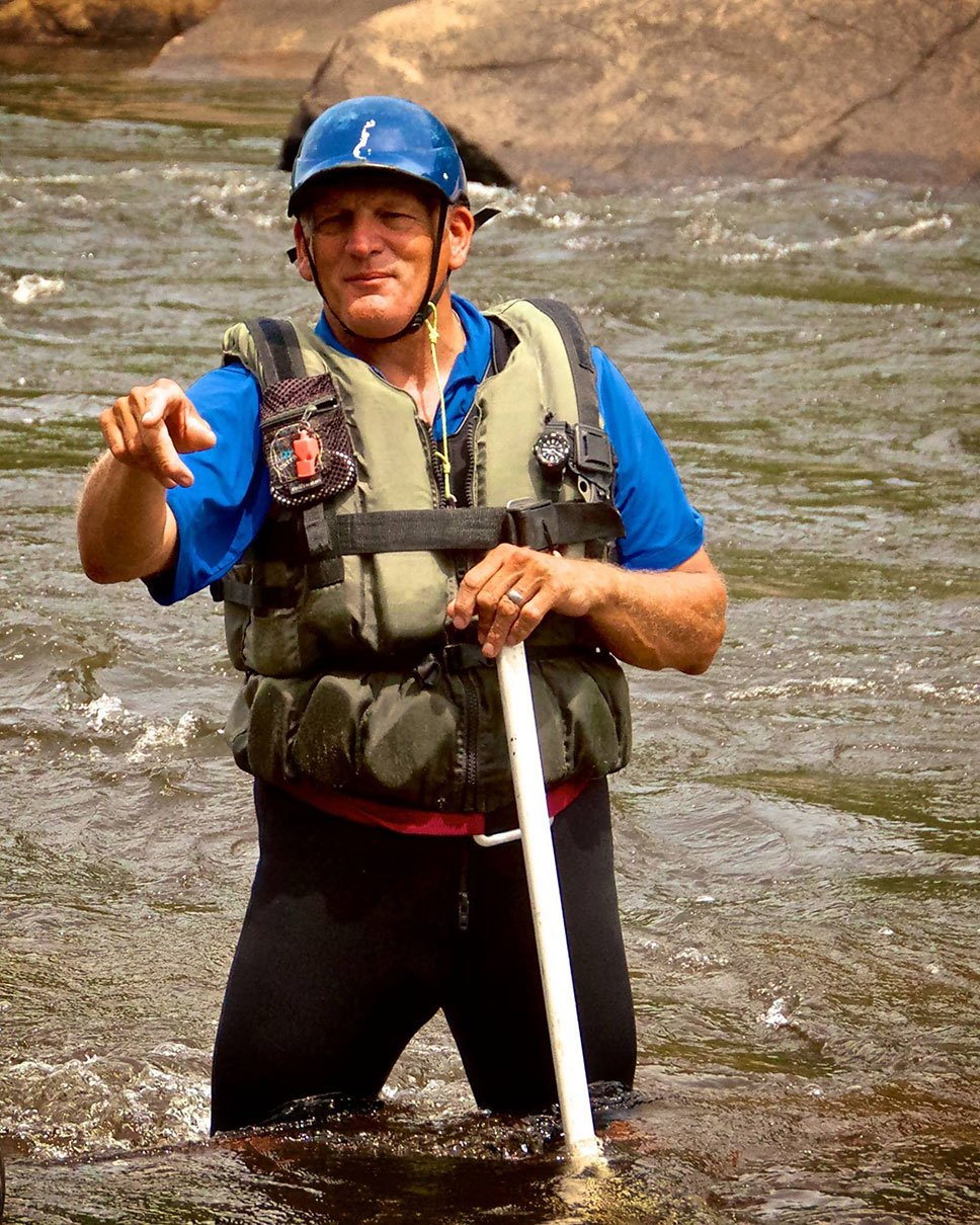 Charlie Walbridge, author of Whitewater Rescues, stands and points in a knee-deep river with rescue gear and paddle