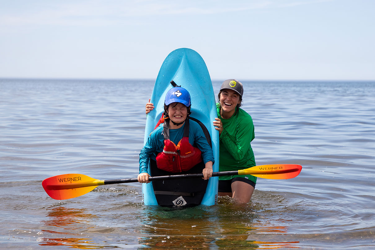 woman instructor smiles as she holds up youth kayak with grimacing teen in it