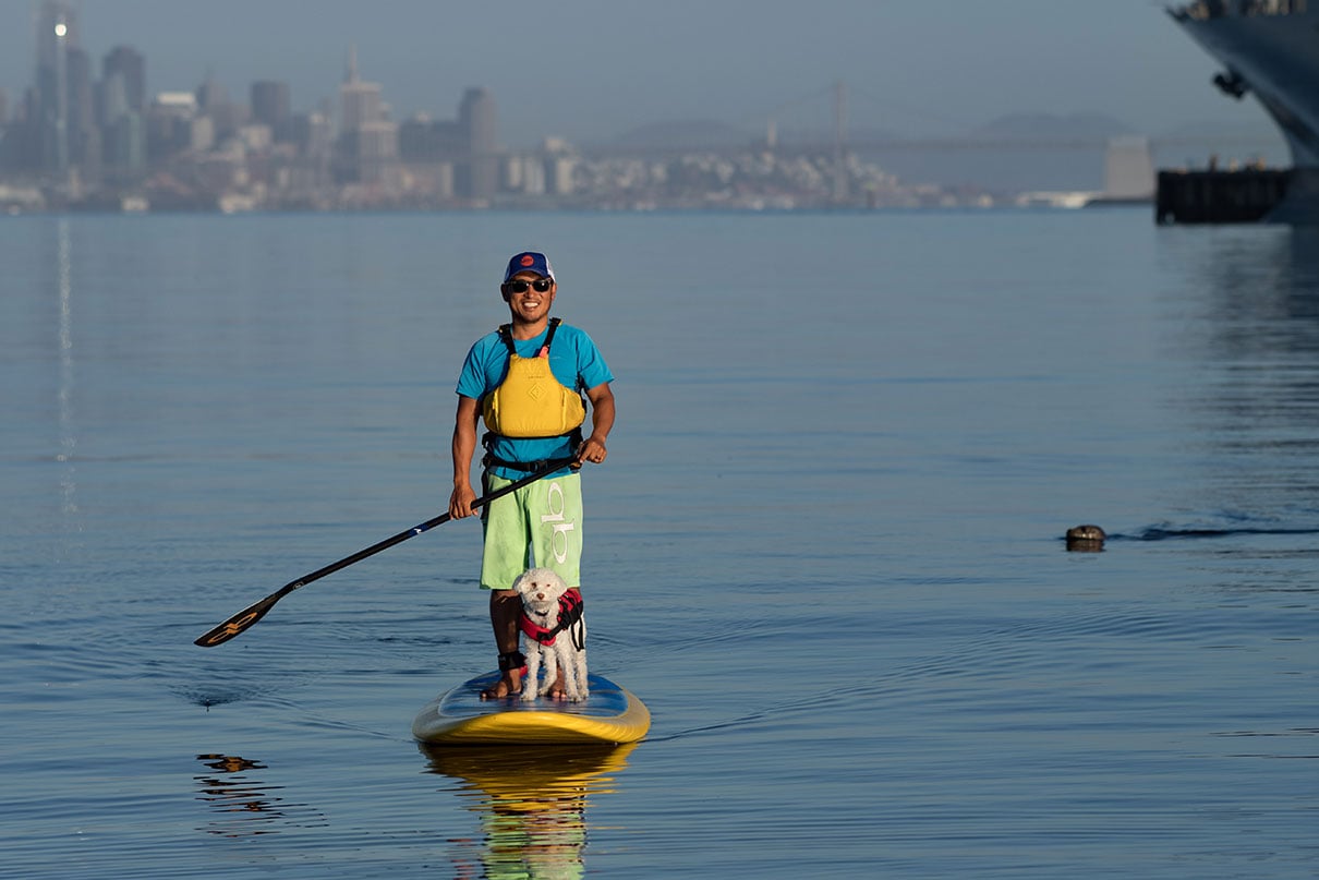 man smiles as he stands on a paddleboard with city skyline in background