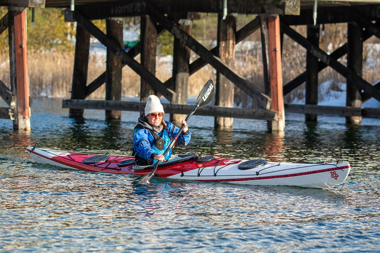 woman paddles a sea kayak past dock pilings in cool weather