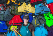 Paddling Magazine editor-in-chief Kaydi Pyette holds a canoe paddle while wearing and surrounded by a selection of her picks for best life jackets