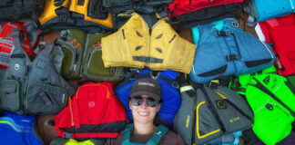 Paddling Magazine editor-in-chief Kaydi Pyette holds a canoe paddle while wearing and surrounded by a selection of her picks for best life jackets