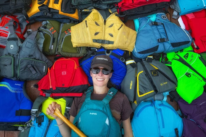 Paddling Magazine editor-in-chief Kaydi Pyette holds a canoe paddle while wearing and surrounded by a selection of her picks for best life jackets