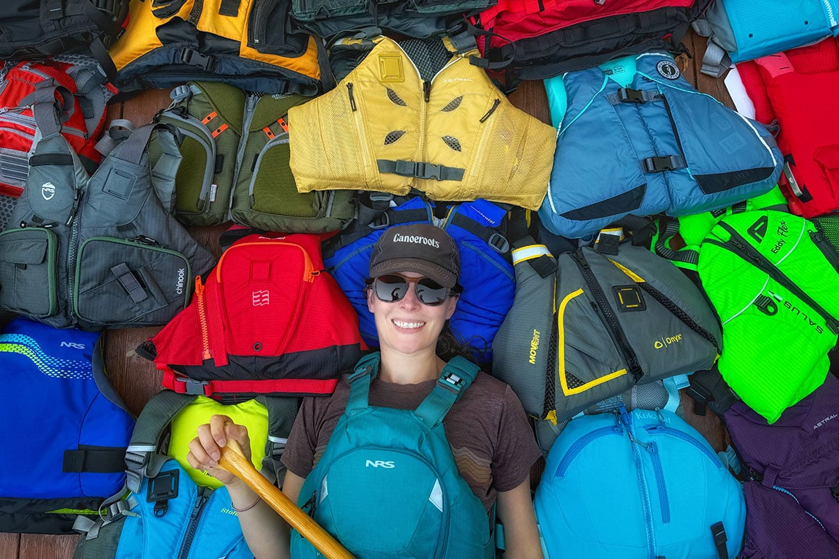 Paddling Magazine editor-in-chief Kaydi Pyette holds a canoe paddle while wearing and surrounded by a selection of her picks for best life jackets