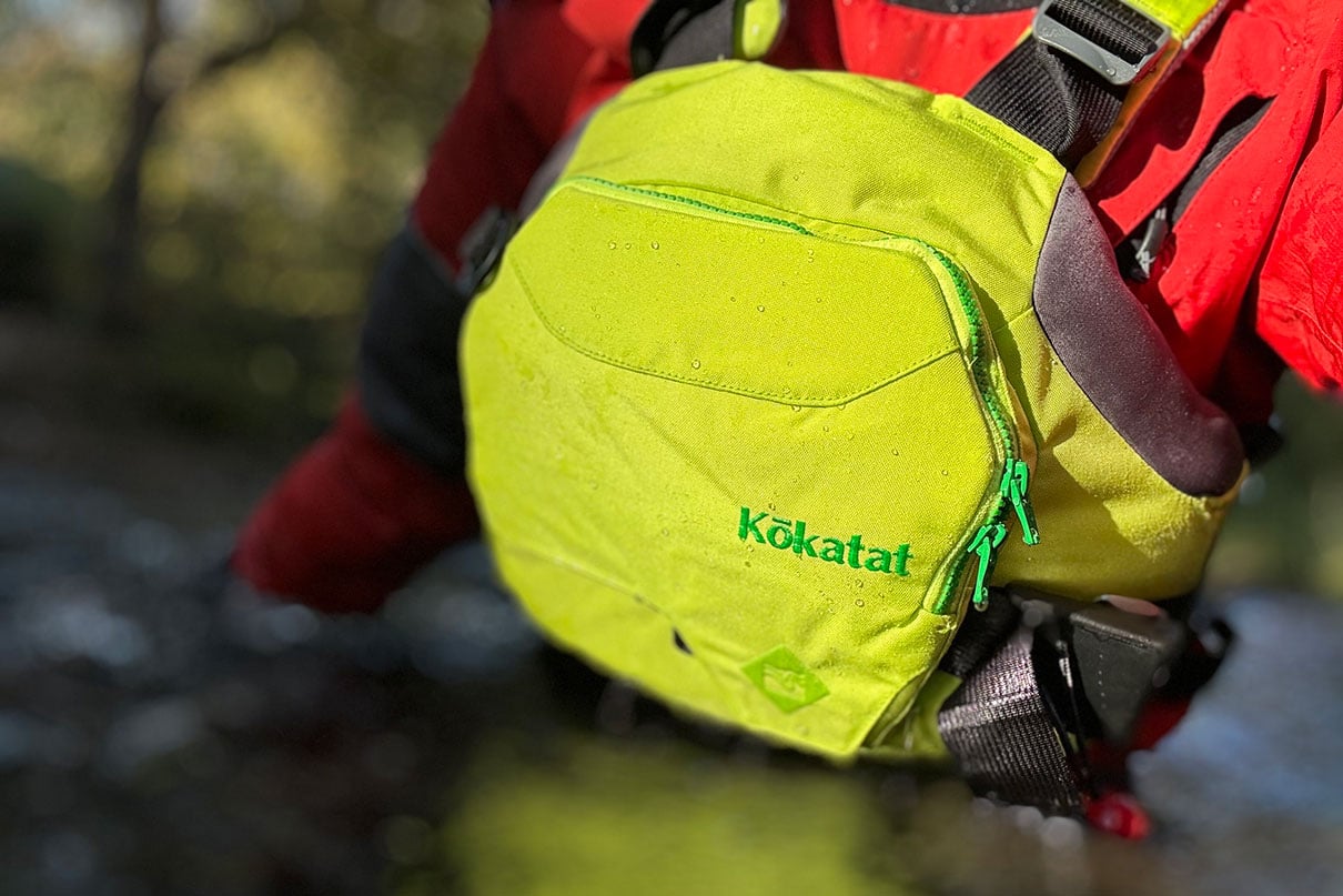 a paddler stands waist-deep in water while wearing red drysuit and yellow Kokatat HustlerR rescue vest