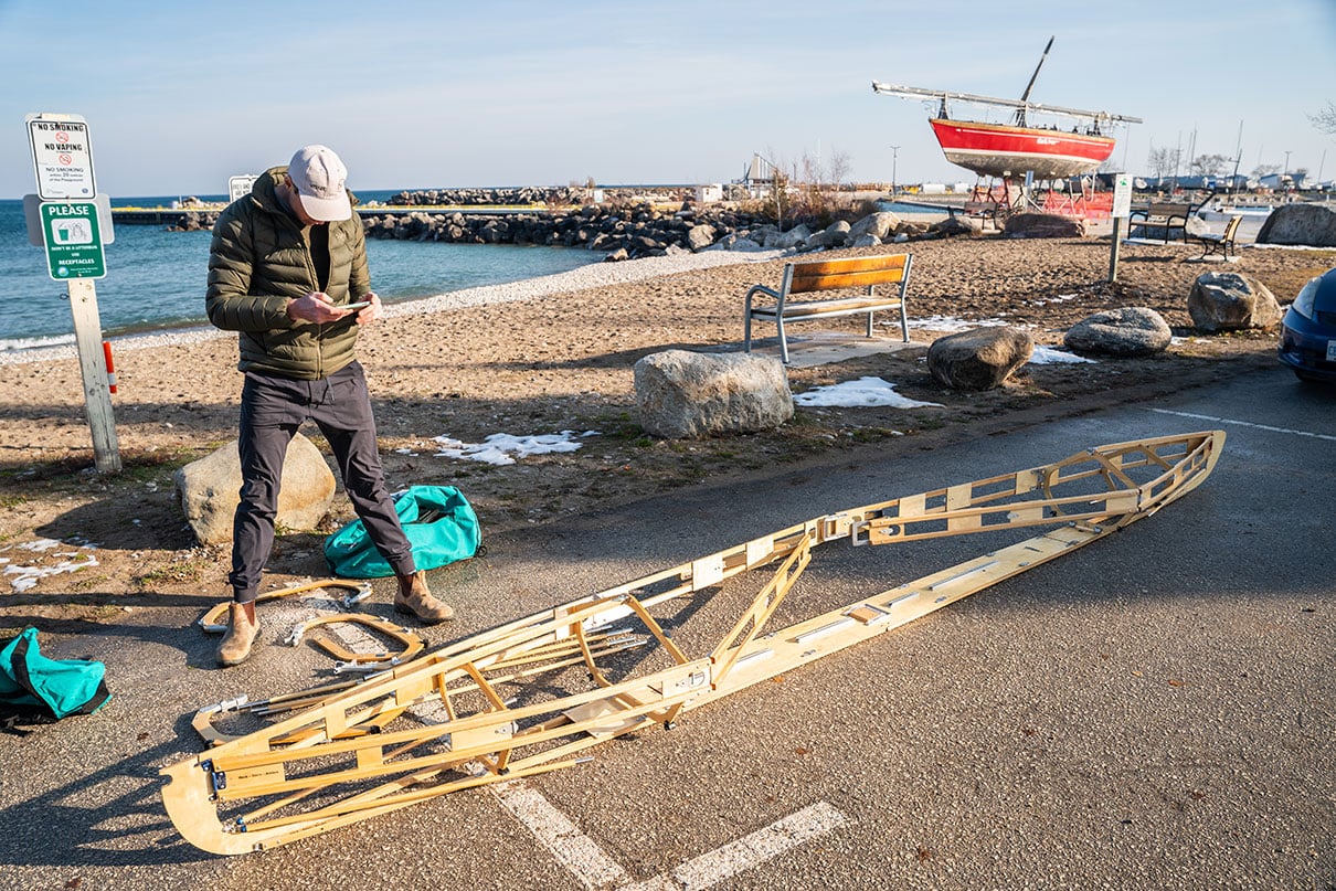 man stands beside the frame of a Klepper while reading instructions at the boat launch