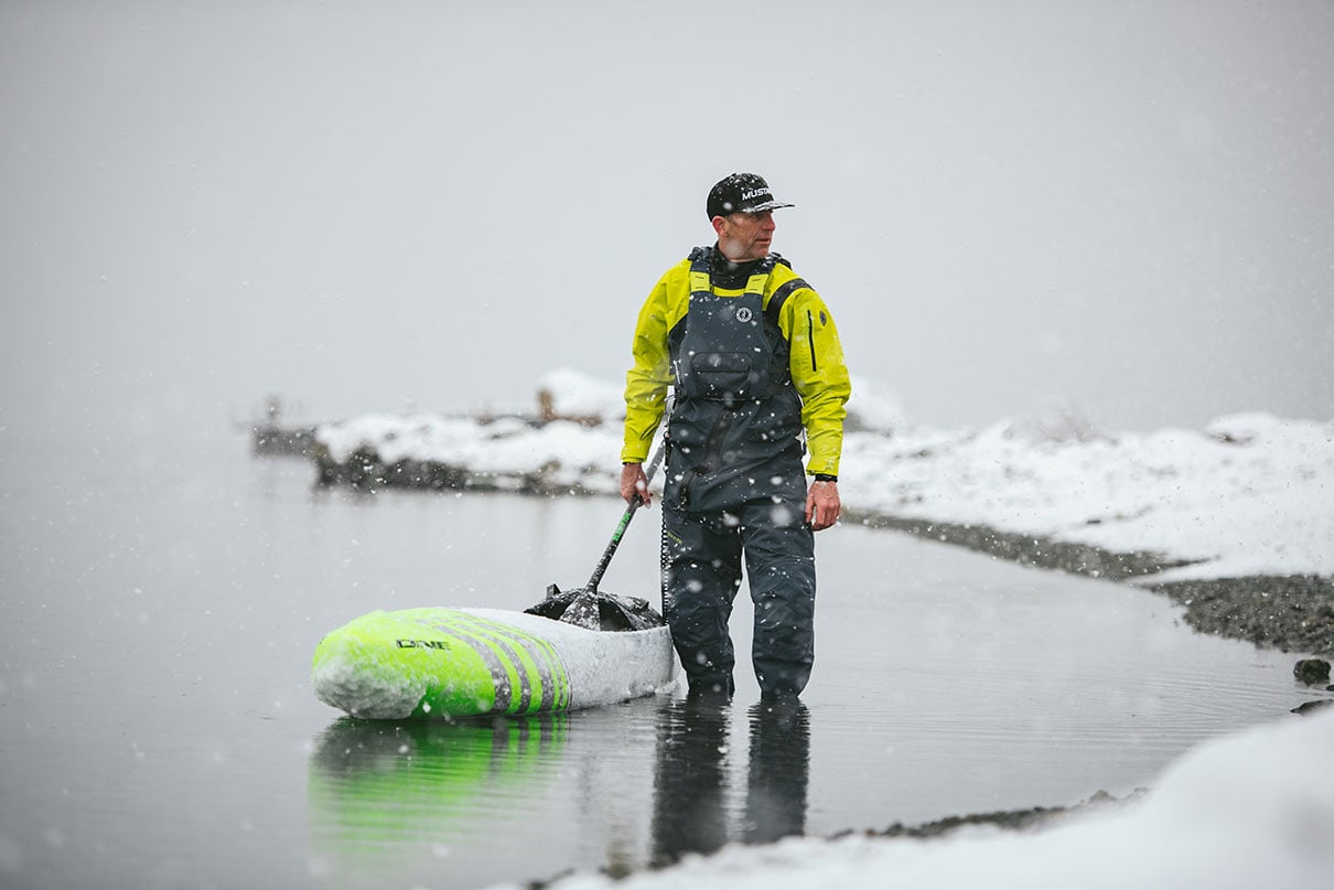 man stands in shallow water with kayak along snowy winter shoreline wearing rescue gear and Khimera PFD