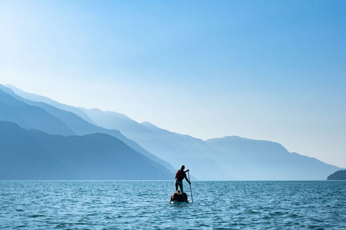 man stands and paddleboards on ocean waters with misty mountains in the background