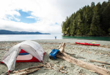 person stands on beach with arms outstretched at remote coastal campsite with kayak, tent and pack nearby