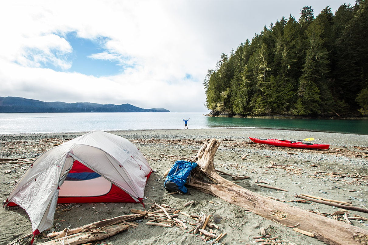 person stands on beach with arms outstretched at remote coastal campsite with kayak, tent and pack nearby