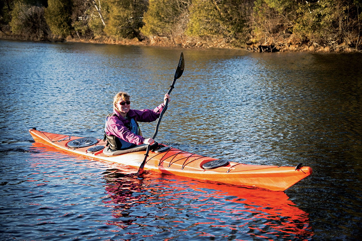 woman paddles the P&H Sea Kayaks Scorpio II touring kayak in front of autumn shoreline