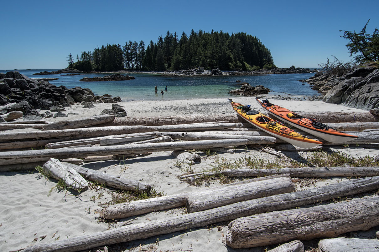 two sea kayaks sit on logs on a remote British Columbia beach while people wade in the shallow water