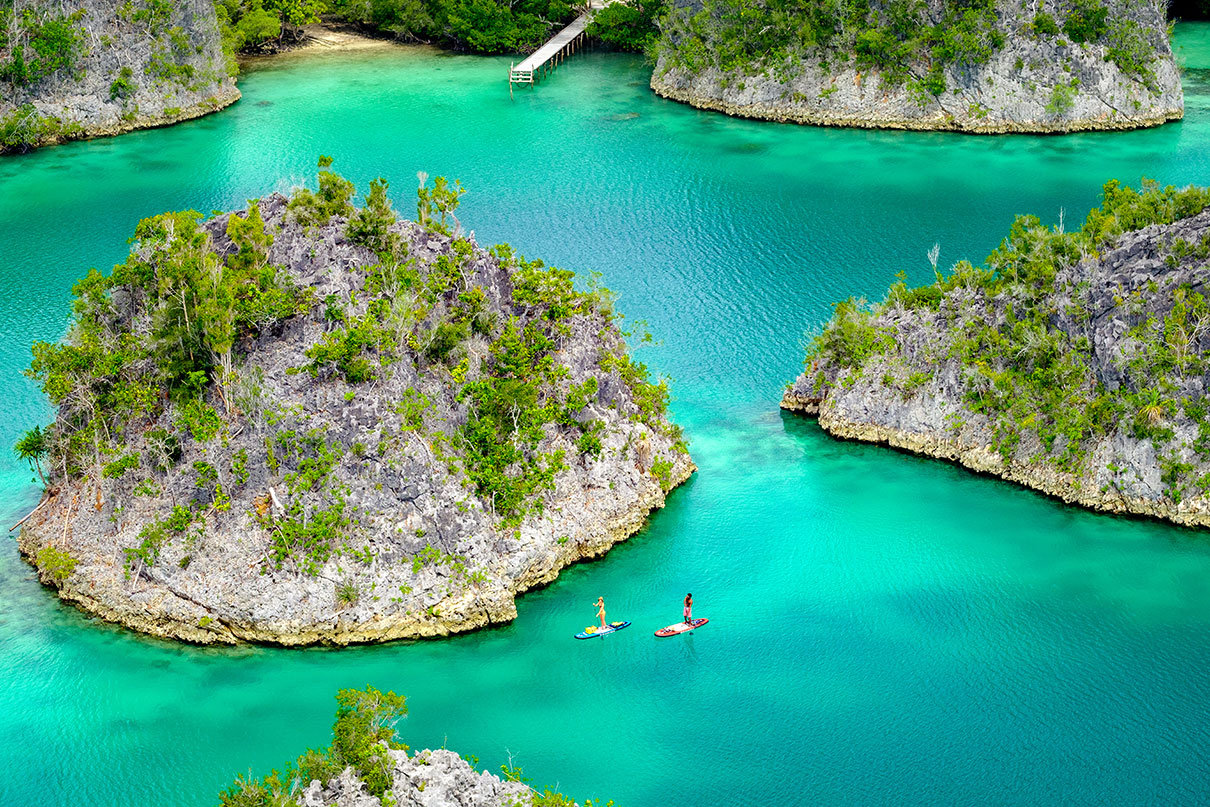 two people paddleboard past small rocky islands in turquoise waters in Indonesia