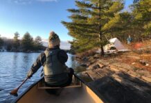 Person dressed in cold weather apparel paddling a canoe toward camp.