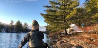 Person dressed in cold weather apparel paddling a canoe toward camp.