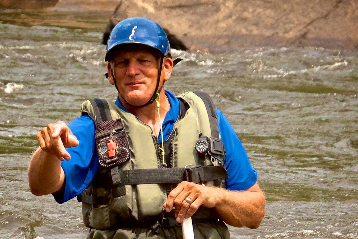 Charlie Walbridge, author of Whitewater Rescues, stands and points in a knee-deep river with rescue gear and paddle